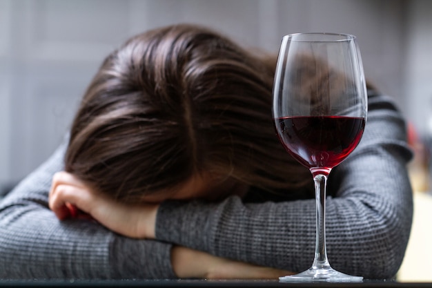 Depressed, divorced crying woman sitting alone in kitchen at home and drinking a glass of red wine because of problems at work and troubles in relationships. social and life problems