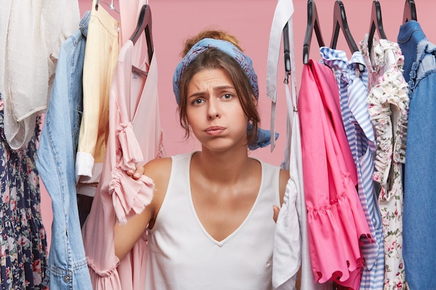 Donna depressa in piedi vicino al guardaroba pieno di vestiti, avendo una scelta difficile non sapere cosa indossare.