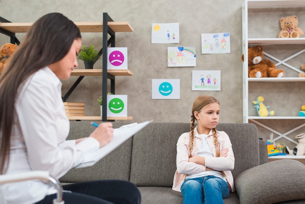 Depressed girl sitting on sofa with female psychologist writing note on clipboard