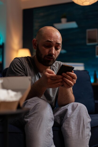 Депрессивный мужчина использует смартфон для просмотра интернета дома, пытаясь вылечить психическое заболевание и тревогу. Грустный отчаявшийся человек с мобильным телефоном в одиночестве, в депрессии.