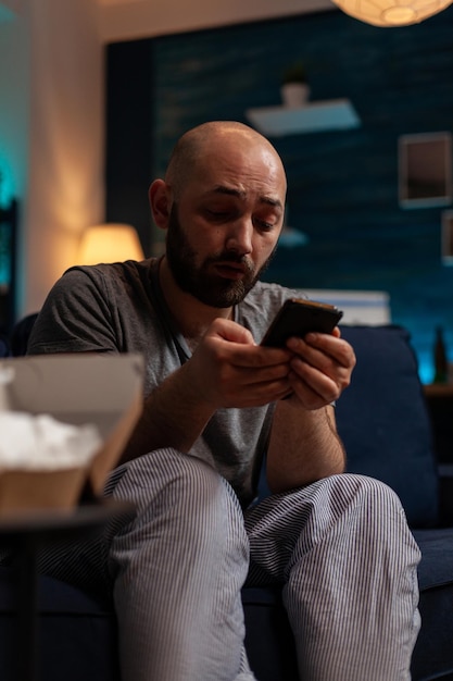 Депрессивный мужчина использует смартфон для просмотра интернета дома, пытаясь вылечить психическое заболевание и тревогу. Грустный отчаявшийся человек с мобильным телефоном в одиночестве, в депрессии.