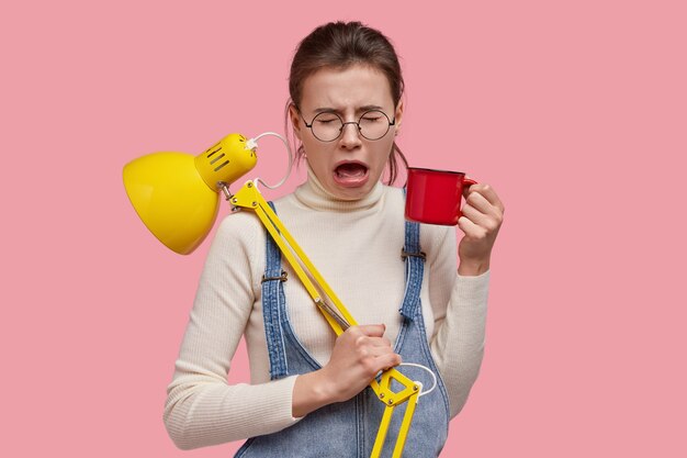 depressed sad woman opens mouth, cries desperately, holds red cup of tea, desk lamp