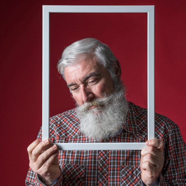 Бесплатное фото Подавленный старший мужчина держит белую рамку перед его лицом на цветном фоне