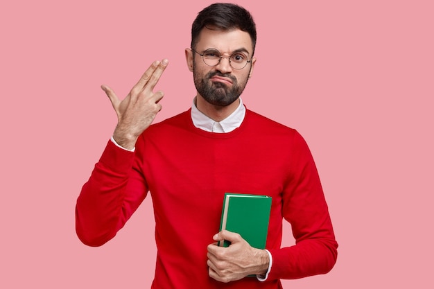 Depressed unshaven male shoots himself in temple, frowns face with displeasure, carries green textbook, dressed in bright red clothes, models over pink space