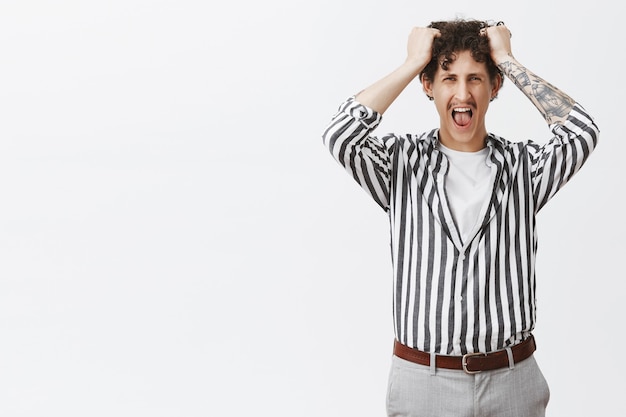 Giovane depresso con i baffi in camicia a righe che perde la pazienza urlando ad alta voce tirandosi fuori i capelli dalla testa stufo e incazzato da uno stupido capo in posa su un muro grigio