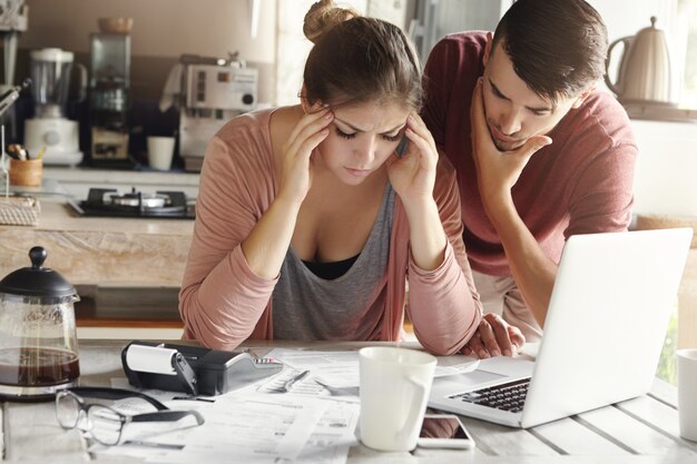 Depressed young woman having bad headache holding her temples feeling stressed by lack of money to pay out family debts, her puzzled husband standing next to her, trying to come up with solution