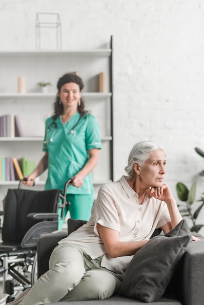 Depressed young woman sitting on sofa in front of nurse standing with wheel chair