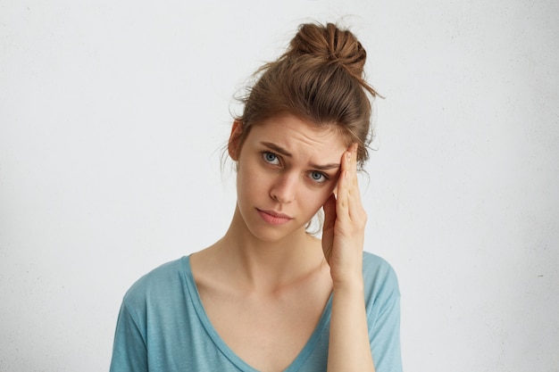 Depressing doleful woman with hair knot, blue exhausted eyes touching her head having discontented expression being tired