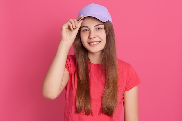 赤いカジュアルなTシャツと野球帽を身に着けている、気分が良く、キャップバイザーに指を置いたまま笑顔の望ましい若い学生の女の子