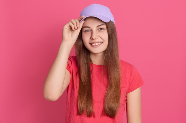 Desirable young smiling student girl wearing red casual t shirt and baseball cap, being in good mood, keeping fingers on cap visor