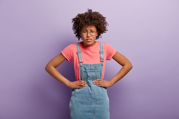 Desperate dark skinned woman looks with upset facial expression, keeps hands on waist, smirks face, wears jean sarafan, argues with someone, feels pain, stands alone against violet wall