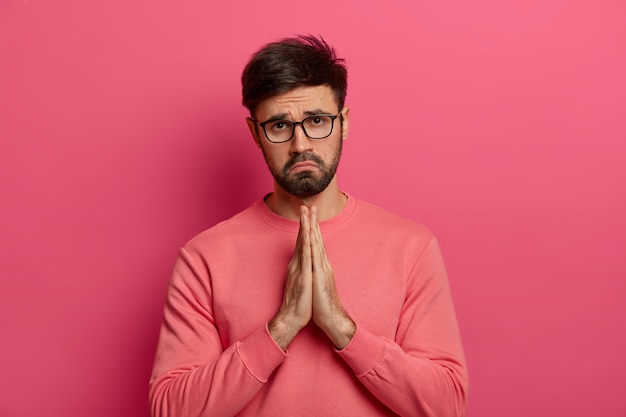 Free photo desperate guilty unshaven man shows clasped hands, says sorry for bad mistake, beggs for help, looks unhappily , wears spectacles and casual jumper, poses indoor over rosy wall.