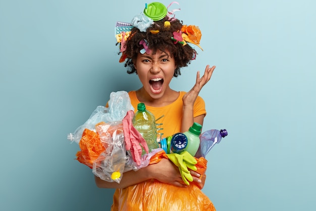 Desperate young Afro American woman fed up of pollution, cleans up rubbish, fights against plastic contamination, dressed in casual t shirt, gestures angrily, isolated against blue wall.