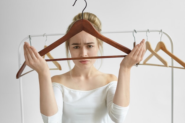 Giovane donna disperata con una crocchia di capelli che ha un'espressione facciale sconvolta, guardando attraverso la gruccia vuota, sentendosi frustrata, non ha vestiti o soldi per comprare un vestito nuovo per un'occasione speciale