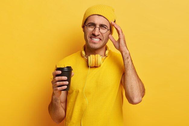 Desperate young man has headache, feels overworked and touches temple, clenches teeth, wears yellow vivid clothes, holds takeaway coffee cup, wears round spectacles. Negative feelings concept