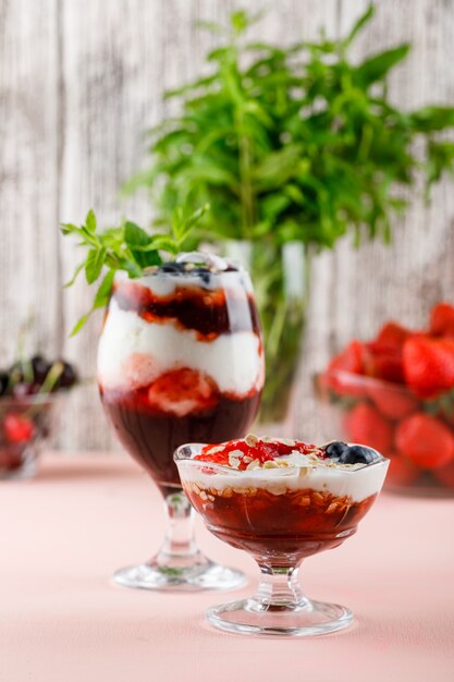 Десерт с клубникой, черникой, мятой, вишней в вазе и бокалом на розовой и шероховатой стене, вид сбоку.