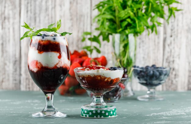 Десерт с клубникой, черникой, мятой, вишней в вазе и бокалом на штукатурке и шероховатой поверхности, вид сбоку.