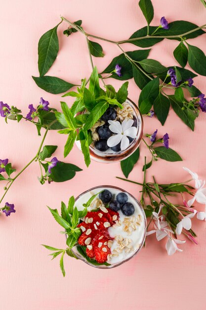 Dessert with strawberries, blueberries, nuts, mint, flower branches in goblet and vase on pink surface, top view.