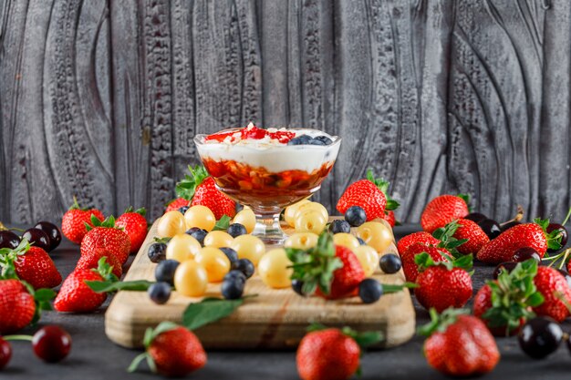 Dessert with strawberry, blueberry, cherry, cutting board in a vase on grey and wooden surface, side view.