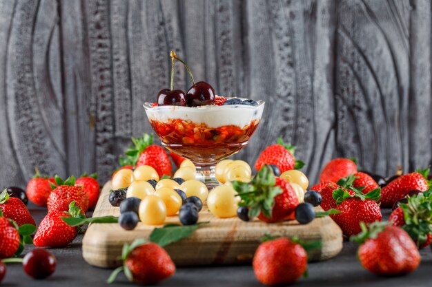 Dessert with strawberry, blueberry, cherry, cutting board in a vase on grey and wooden surface, side view.