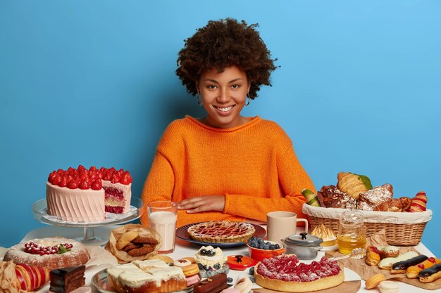 Desserts, fast food, unhealthy lifestyle concept. Pleased dark skinned model in orange jumper, enjoys party, has no diet, boosts mood with sweet dishes, isolated on blue wall.