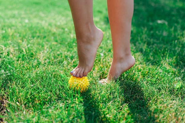 Details of the bare feet of a young woman standing on tiptoes on a spiky rubber yellow ball to relax...