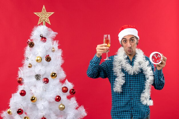 Determined emotional young man with santa claus hat and holding a glass of wine and clock near Christmas tree on red