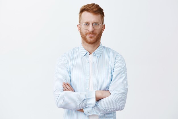 Determined good-looking neat male redhead in glasses and shirt, holding hand crossed on chest in self-assured posture and looking, listening carefully