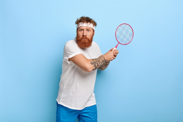 Determined red haired tennis player holds racket while posing against the blue wall