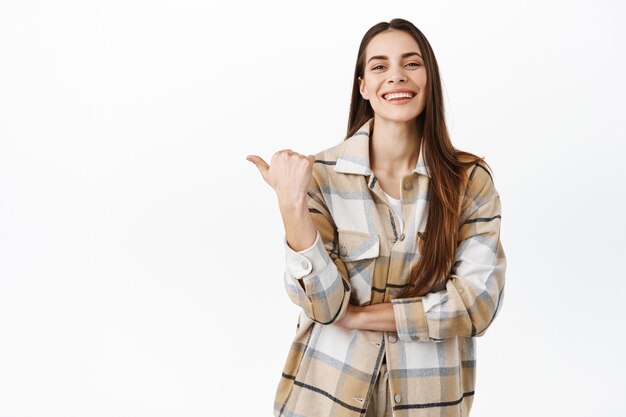 Determined smiling woman pointing aside at copyspace, showing left side logo and looking pleased and happy at front, standing over white wall