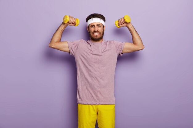 Determined young sporty unshaven man clenches teeth, raises muscular arms, does exercises with dumbbells, clenches teeth