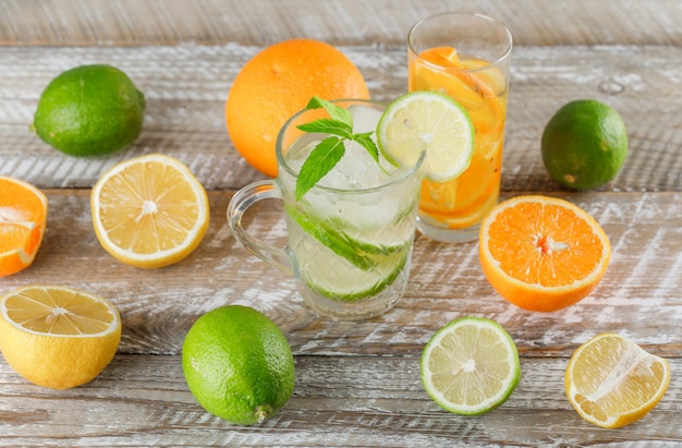 Detox water with limes, lemons, oranges, mint in cup and glass on wooden surface, high angle view.