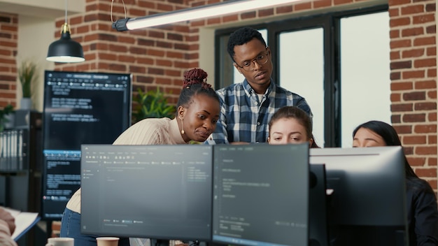 Developers brainstorming ideas looking at code on computer screens asking feedback from senior developer while intern joins the discussion. Junior programmers collaborating on group project.