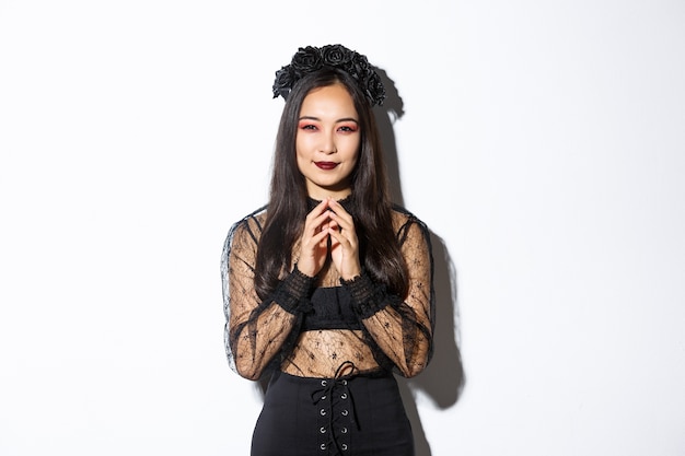 Devious young asian woman in gothic lace dress smiling pleased and steeple fingers. Witch prepare evil plan, smiling cunning and looking at camera. Halloween concept.