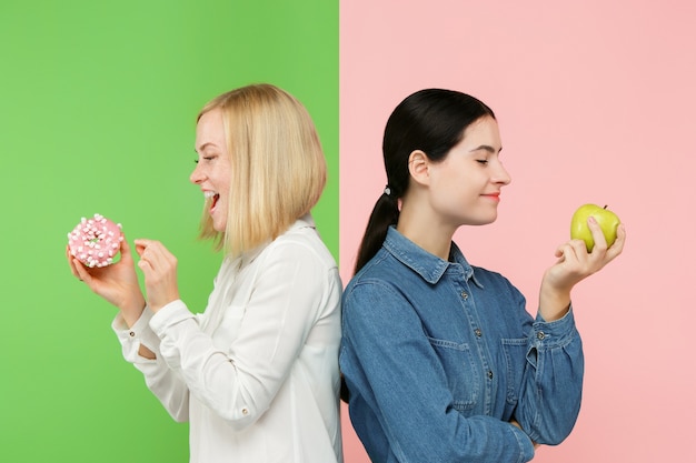 Diet. Dieting concept. Healthy useful food. Beautiful young women choosing between fruits and unhelathy cake at studio. Human emotions and comparison concepts