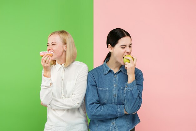 Dieting concept. Healthy useful food. Beautiful young women choosing between fruits and unhealthy cake at studio. Human emotions and comparison concepts