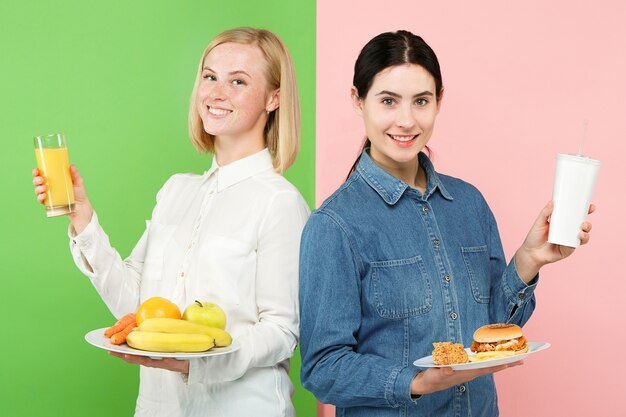 Dieting concept. Healthy useful food. Beautiful young women choosing between fruits and unhealthy fast food at studio. Human emotions and comparison concepts