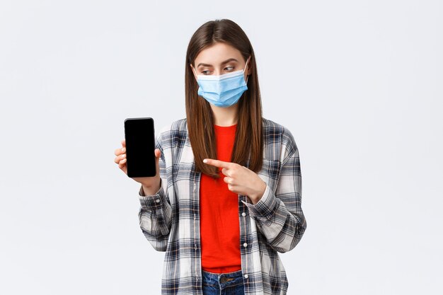 Different emotions, covid-19, social distancing and technology concept. Attractive 20s female in medical mask, pointing finger at mobile phone, advice download app or game, filters for photos