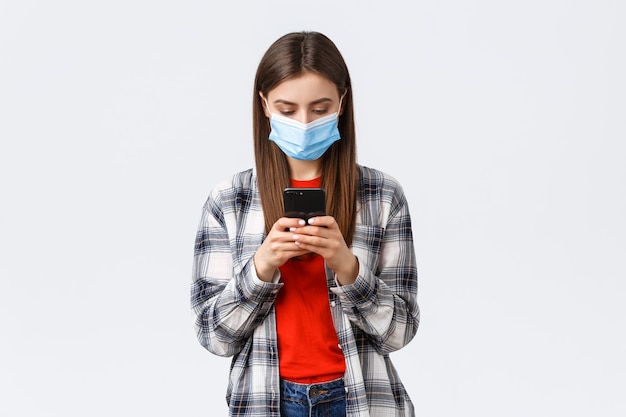 Emozioni diverse, covid-19, distanza sociale e concetto di tecnologia. attraente giovane donna in maschera medica che invia messaggi di testo, guardando lo schermo del telefono cellulare occupato, lavorando da casa