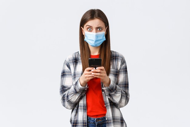 Different emotions, covid-19, social distancing and technology concept. Puzzled and confused young woman in medical mask react to strange message, hold mobile phone, look camera unsure.