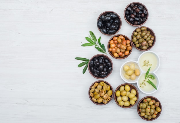 Different kinds of olives and olive oil in a clay and white bowls with olive leaves flat lay on white wood