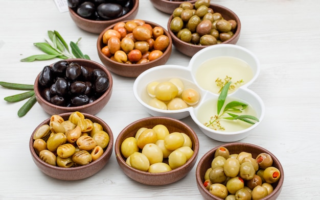 Olive e olio d'oliva differenti in un'argilla e ciotole bianche con la vista dell'angolo alto delle foglie di ulivo su legno bianco