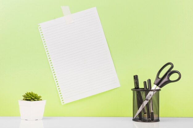 Different pens and scissors in holder near blank paper stucked on wall