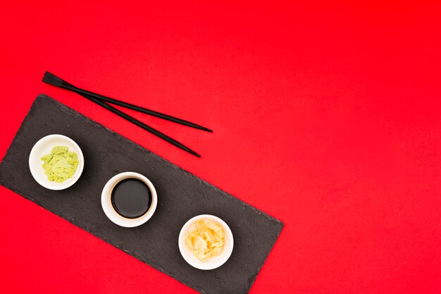 Различные вкусные соусы с маринованным имбирем в мисках и палочками на черном камне над красным столом