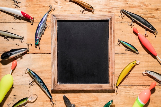 Different type of fishing lures around the black wooden slate on wooden backdrop