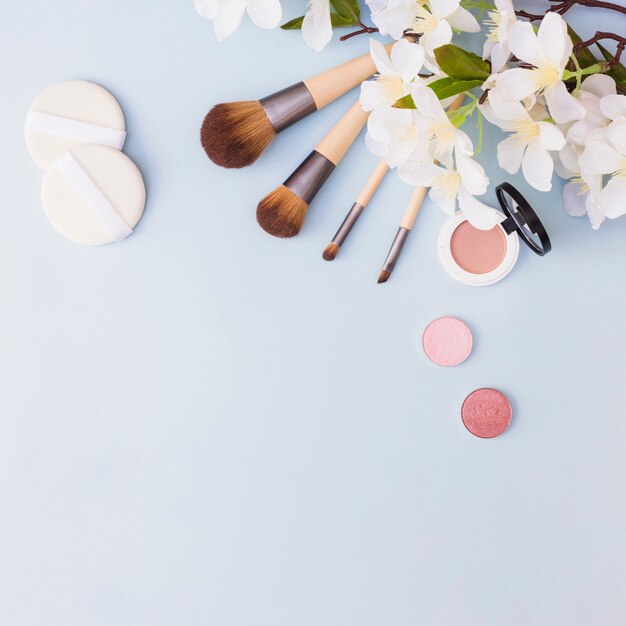 Different type of makeup brush; sponge; eye shadow and blusher with white flowers on blue background