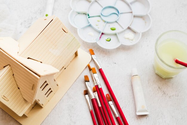 Different Type Of Paintbrush; House Model And Color Palette And Acrylic Paint Tube On White Textured Backdrop