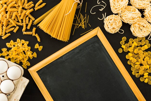 Different type of uncooked pasta with eggs and blackboard on black background