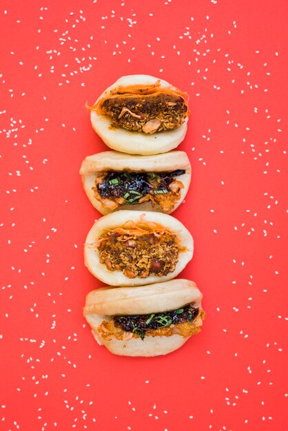 Different types of asian sandwich steamed gua bao buns surrounded with white sesame seeds on red background