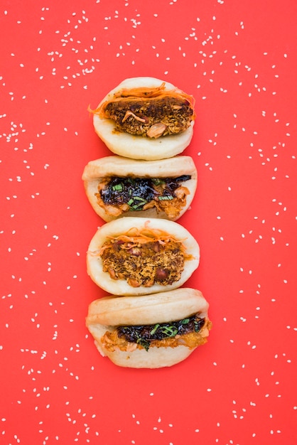 Different types of asian sandwich steamed gua bao buns surrounded with white sesame seeds on red background